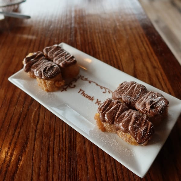 Four churro bites topped with chocolate drizzle and powdered sugar on a white plate with "Thank You" written.