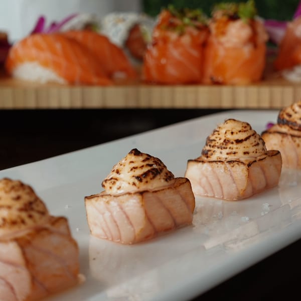 Close-up of seared salmon nigiri with creamy topping on a white plate, with more sushi in the background.