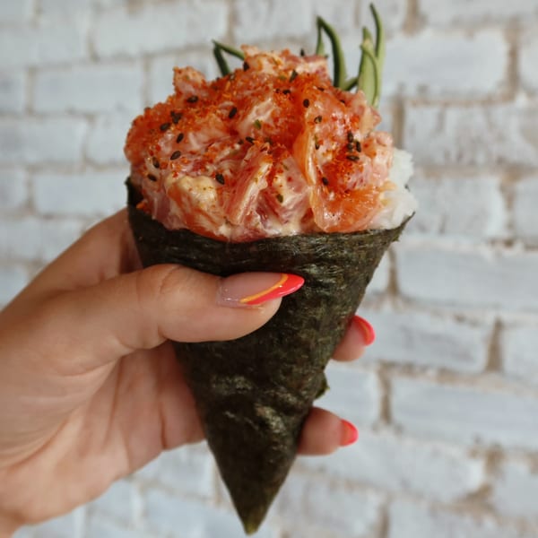 A hand holds a sushi hand roll filled with salmon, rice, and spices against a white brick wall.