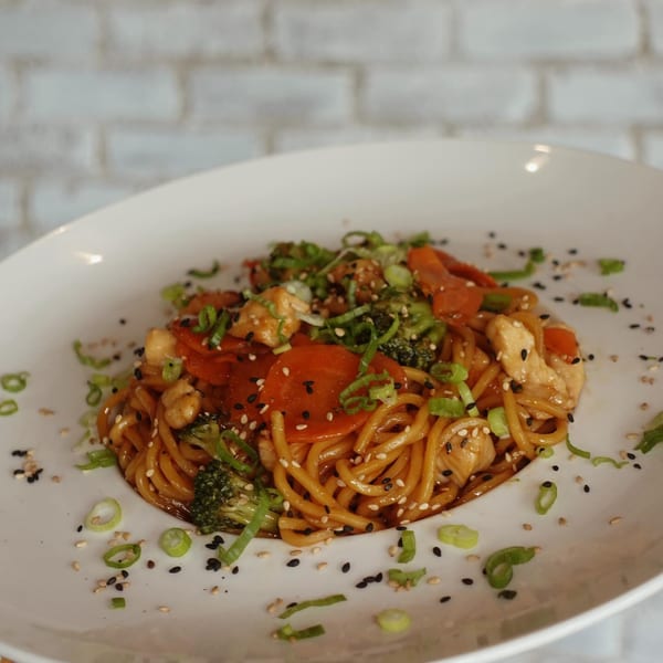 Chicken lo mein with broccoli, carrots, and scallions, garnished with sesame seeds.