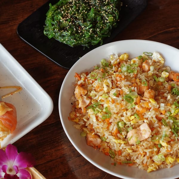 Chicken fried rice, seaweed salad, and salmon sushi on a wooden table with an orchid.