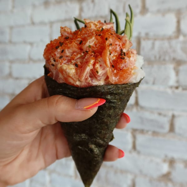 Hand holding a salmon hand roll with spicy mayo, sesame seeds, and chili flakes.