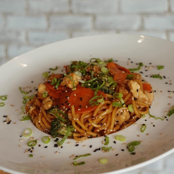 Chicken lo mein with broccoli, carrots, and scallions, garnished with sesame seeds on a white plate.