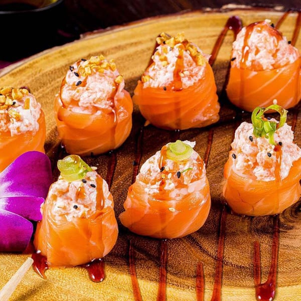 Seven salmon sushi rolls with creamy topping, sesame seeds, sauce, and green garnish on a wooden board.