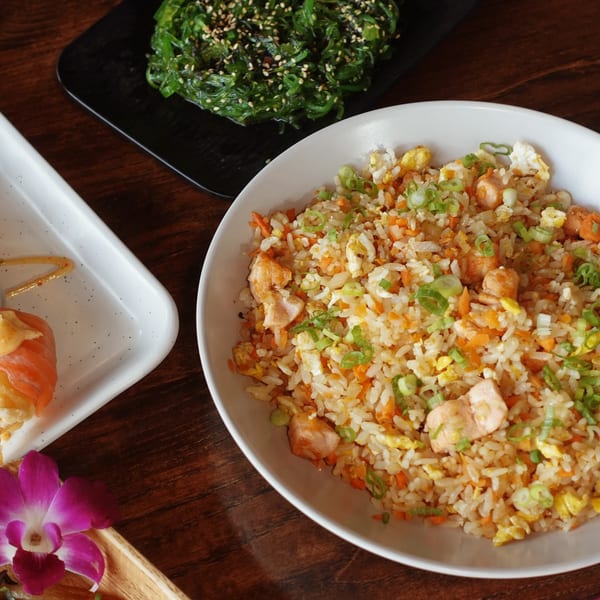 Chicken fried rice, seaweed salad, and sushi on a wooden table with an orchid.