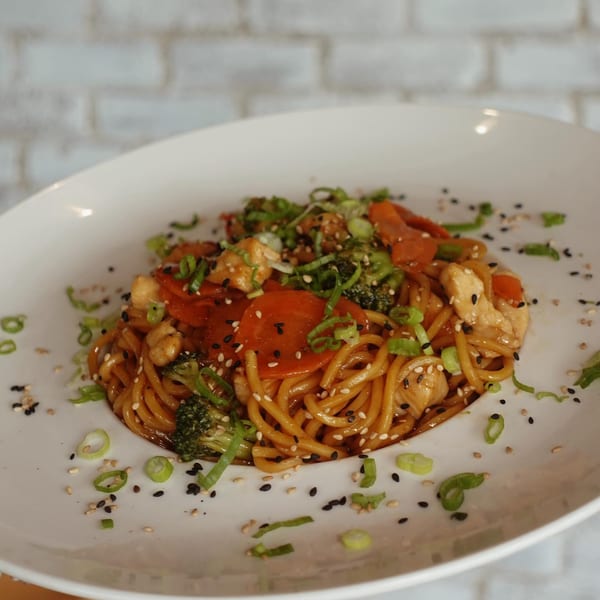 A white plate holds a serving of chicken lo mein with broccoli, carrots, and scallions.