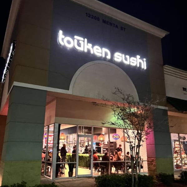 Exterior of Touken Sushi at night, with a glowing sign and patrons visible inside through large windows.