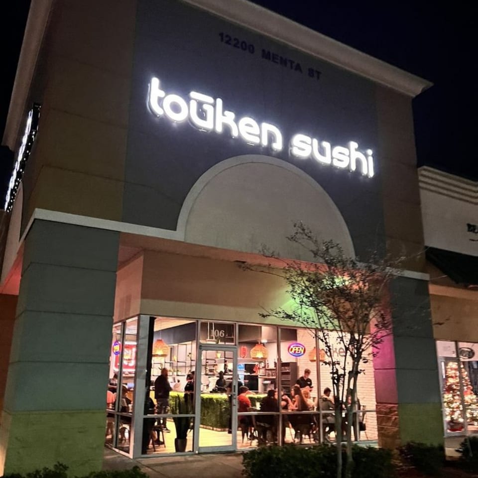 Exterior of Touken Sushi at night, with a glowing sign and patrons visible inside through large windows.
