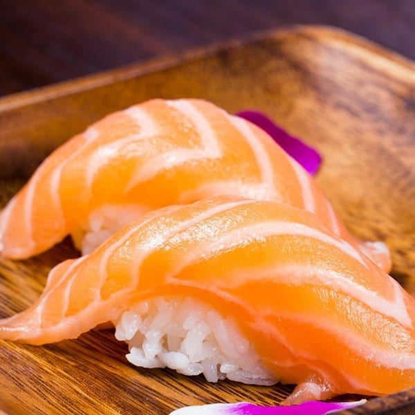 Two pieces of salmon nigiri sushi with white rice on a wooden plate, garnished with purple petals.