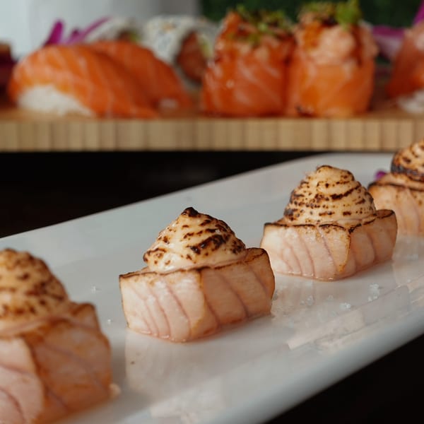 Seared salmon nigiri with creamy topping on a white plate, with more sushi in the background.