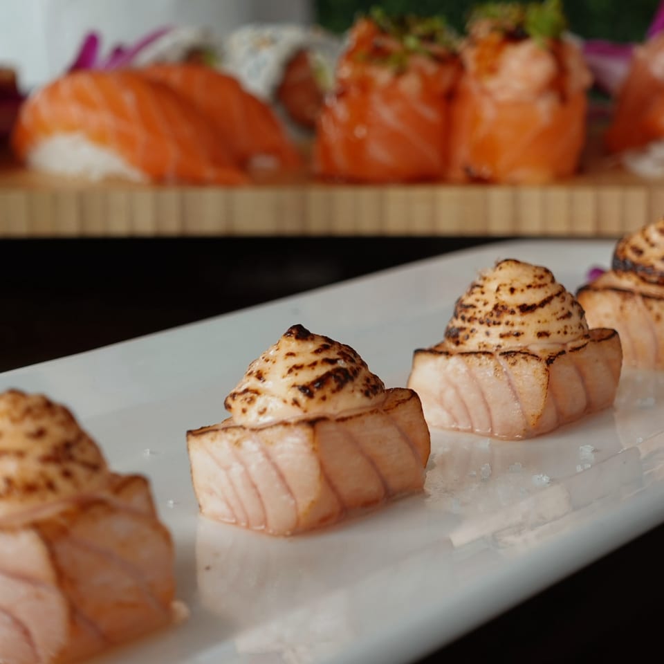 Seared salmon nigiri with creamy topping on a white plate, with more sushi in the background.
