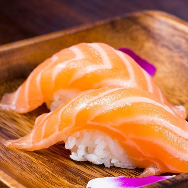 Two pieces of salmon nigiri sushi with white rice on a wooden plate, garnished with purple petals.
