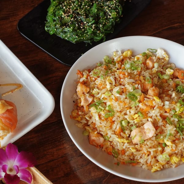 Chicken fried rice, seaweed salad, and sushi on a wooden table with an orchid.