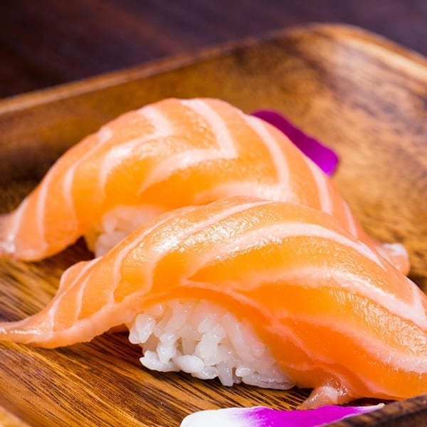 Two pieces of salmon nigiri sushi with white rice on a wooden plate, garnished with purple petals.