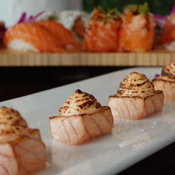 Seared salmon nigiri with creamy, charred topping on a white plate, with more sushi in the background.
