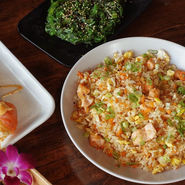A bowl of chicken fried rice, a plate of seaweed salad, and sushi on a wooden table.