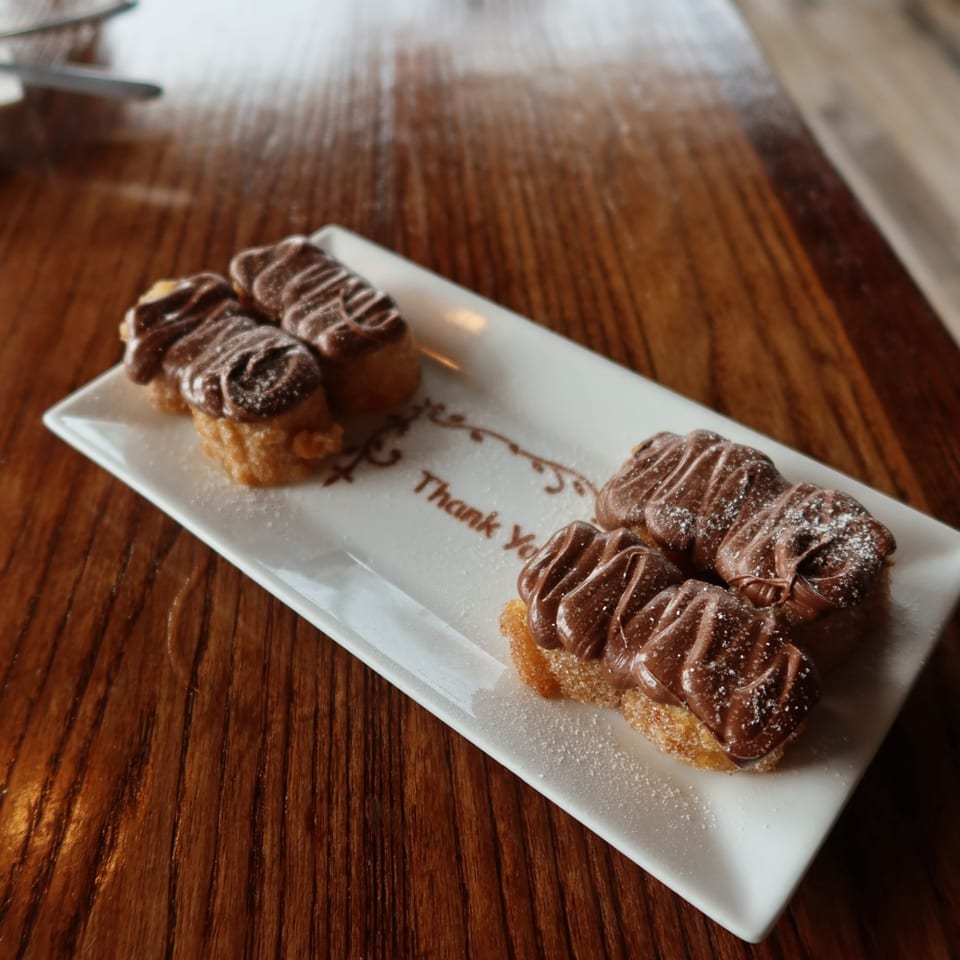 Four churro bites topped with chocolate drizzle and powdered sugar on a white plate with "Thank You" written.