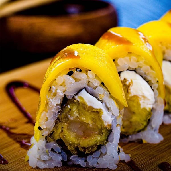 Close-up of three sushi rolls with mango, cream cheese, and tempura shrimp on a wooden board.